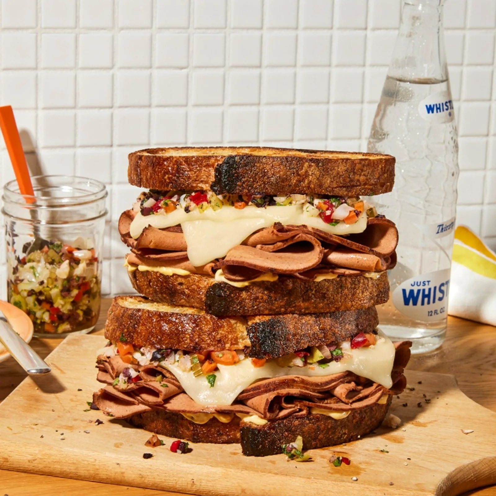 two loaded melty deli sandwiches stacked on top of each other with vegan ham on a wooden cutting board