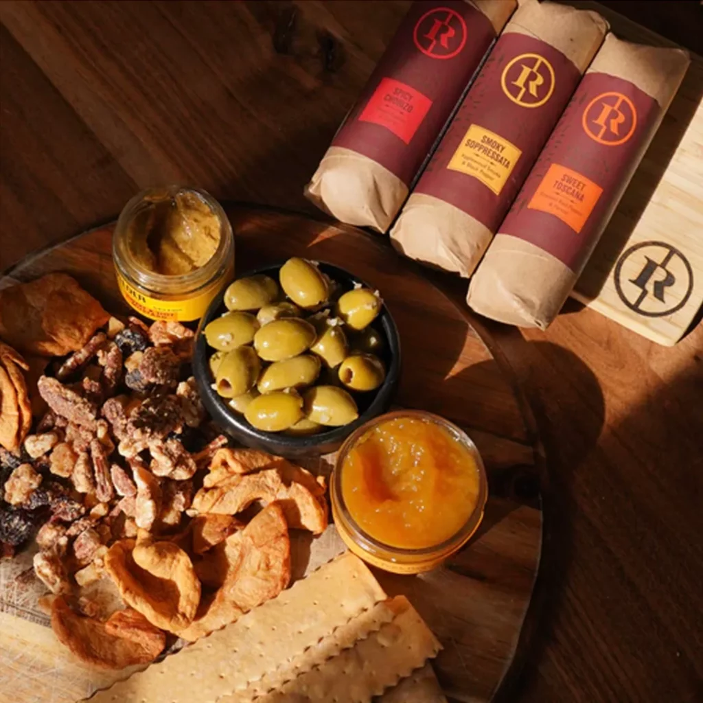 three pack of vegan salami next a serving board of nuts, jam, crackers, and olives