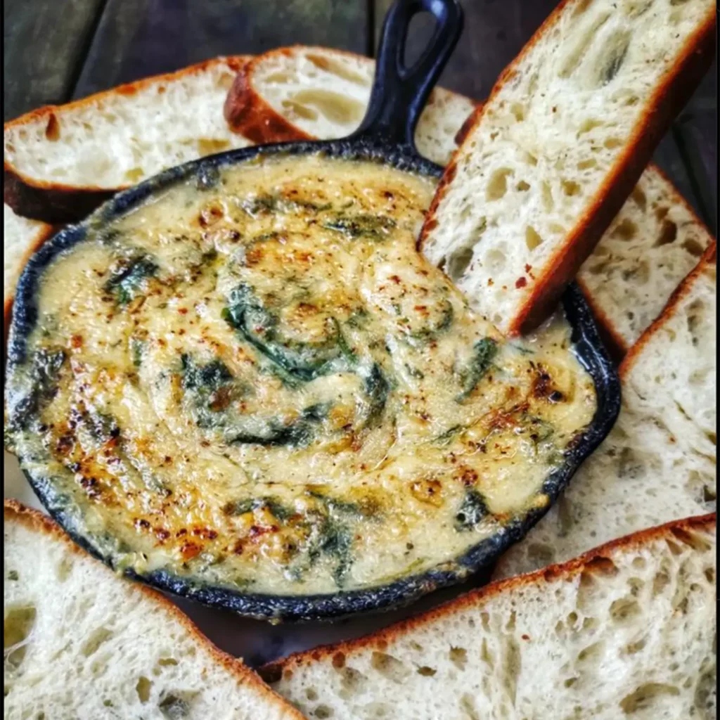 vegan smoked gouda spinach dip in a skillet with bread slices surrounding it and one slice dipped in skillet