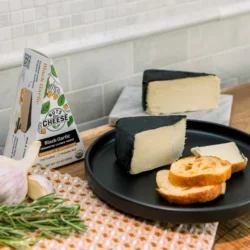 A wedge of vegan black garlic cheese and bread on a black plate, a cloth towel with onion and herbs on a wooden table