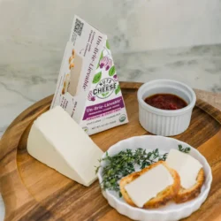 A wedge of vegan brie, a small plate with bread and brie slices, a small bowl of jam on a wooden charcuterie board