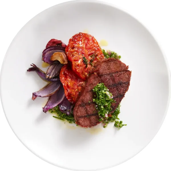Vegan plant-based steak on a white plate with roasted tomatoes onions and pesto by Chunk Foods