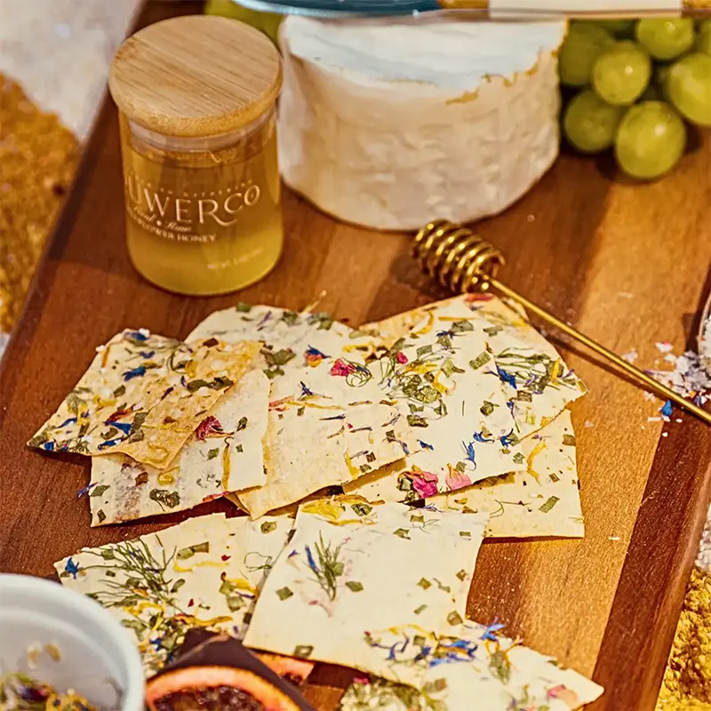 Vegan plant-based crackers made with edible flowers and dried herbs on a board with a wheel of brie and honey