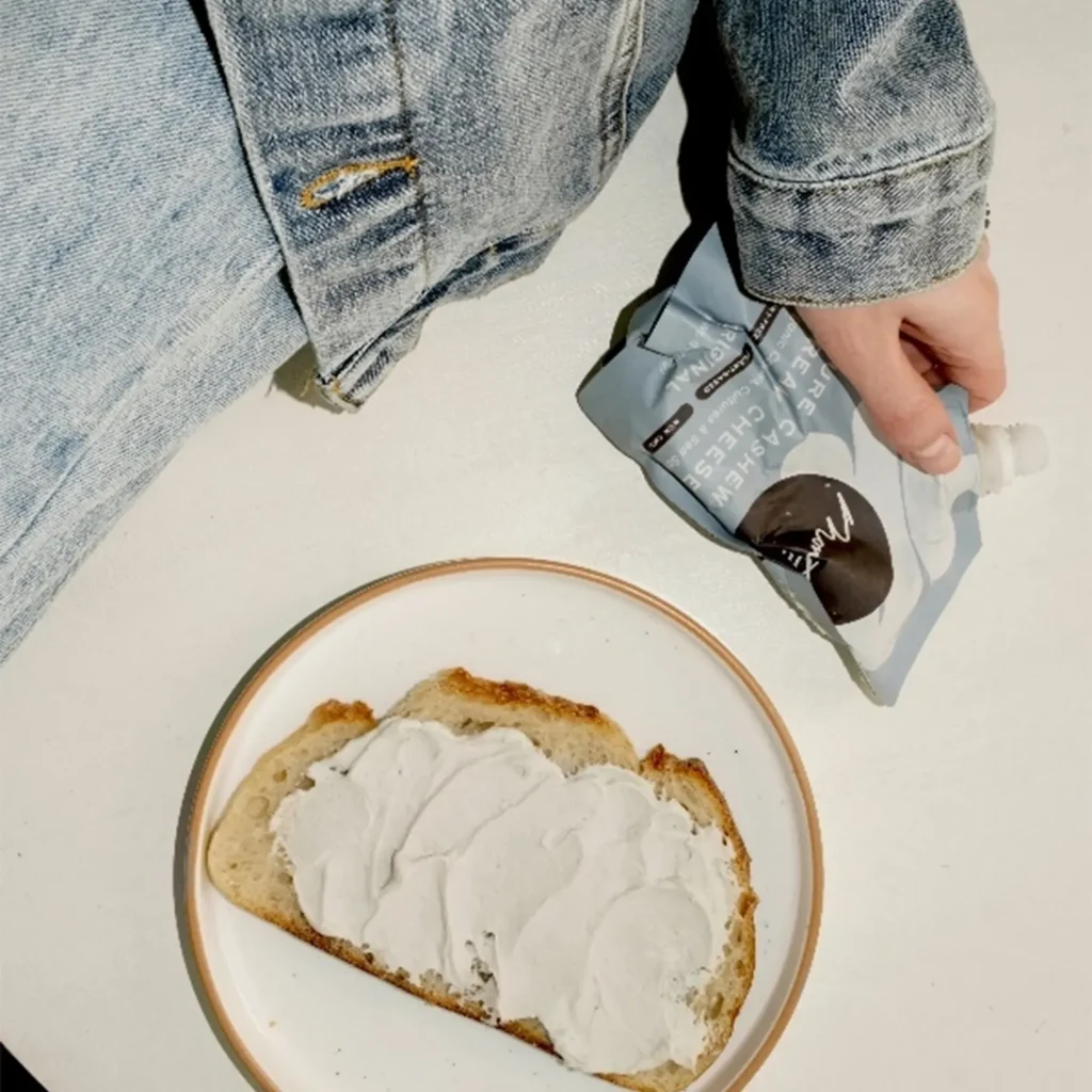 person holding a package of vegan cream cheese sitting on a white table with a plate of bread with cream cheese smeared on it