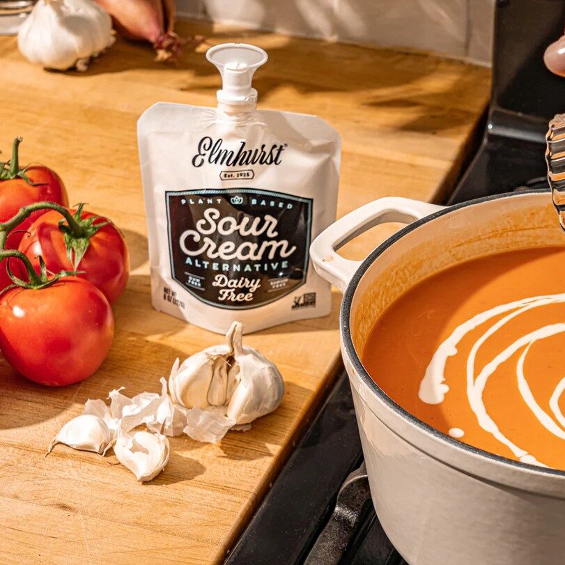 vegan sour cream on a wooden table with tomatoes, garlic and a pot of sauce on a stove that someone is pouring sour cream into.
