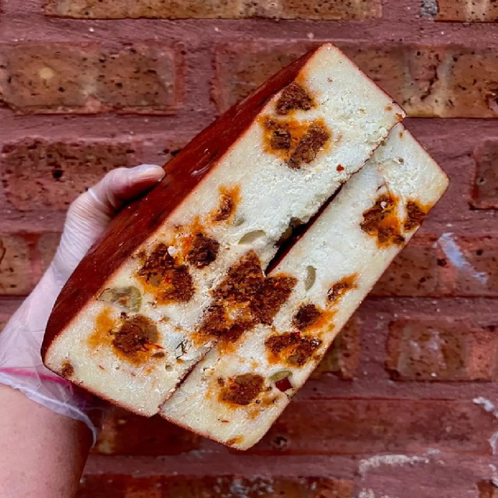 gloved hand holding vegan chorizo manchego cheese wheel cut in half with a brick wall background