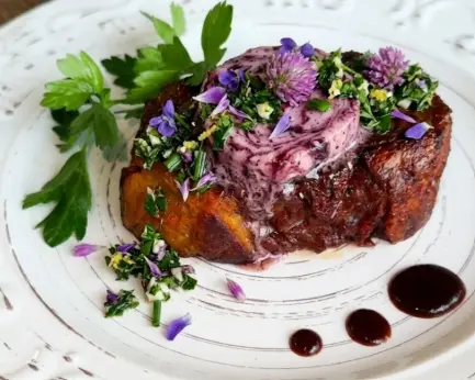 A close-up, top-down shot of a seared and smoked vegan steak, served on a grey artisanal plate, generously topped with melting purple Port Wine compound butter and a vibrant wildflower gremolata with edible purple and yellow petals.