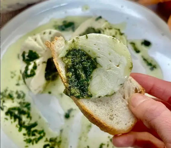 A close-up shot of a hand dipping a piece of fresh, crusty bread into a vibrant, homemade green basil pesto served in a small white bowl.