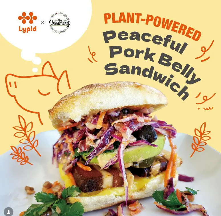 A gourmet, plant-based bacon banh mi sandwich cut in half, showcasing layers of thick-cut Lypid bacon, quick-pickled carrots and cucumbers, fresh cilantro sprigs, jalapeno slices, and a spicy vegan mayo drizzle on a toasted baguette.
